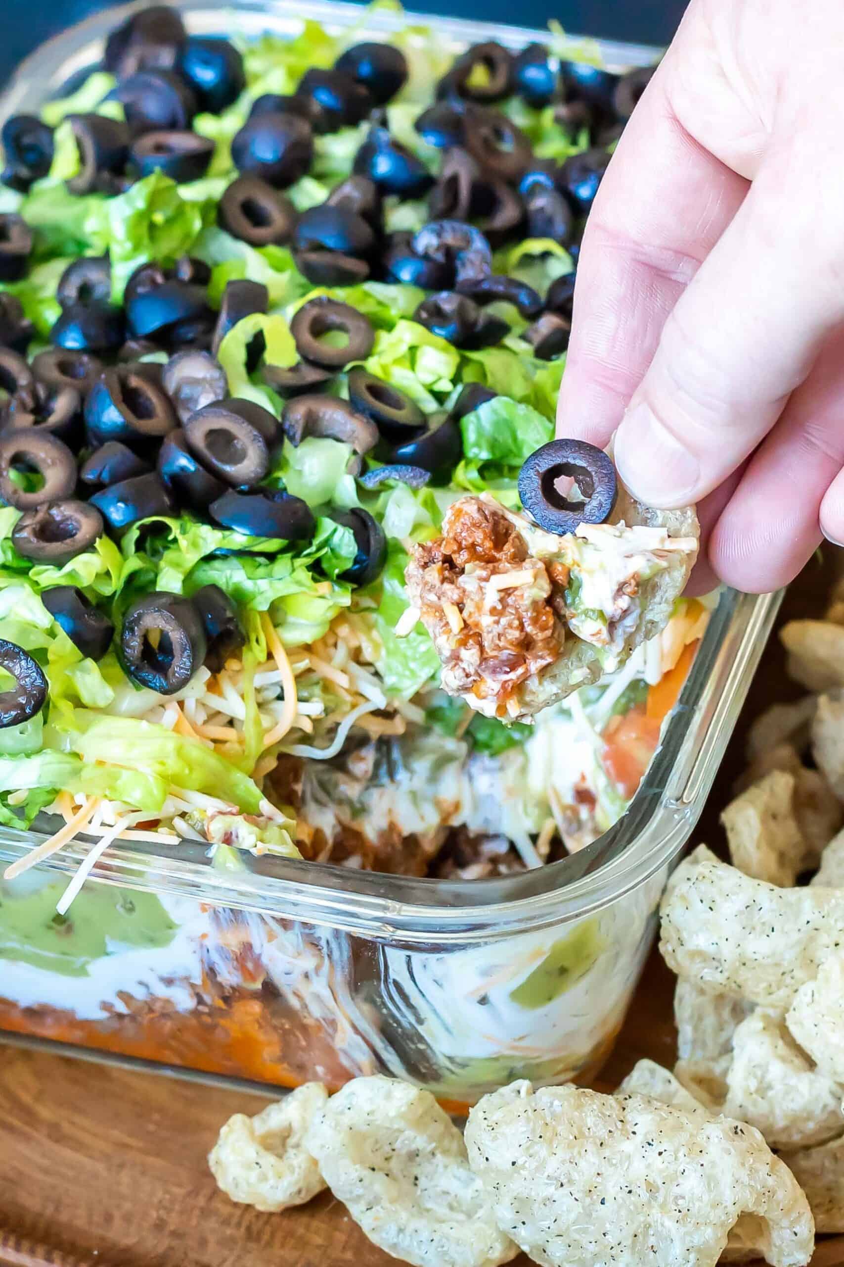 A hand taking a scoop out of the Keto 7 Layer Taco Dip with a pork rind